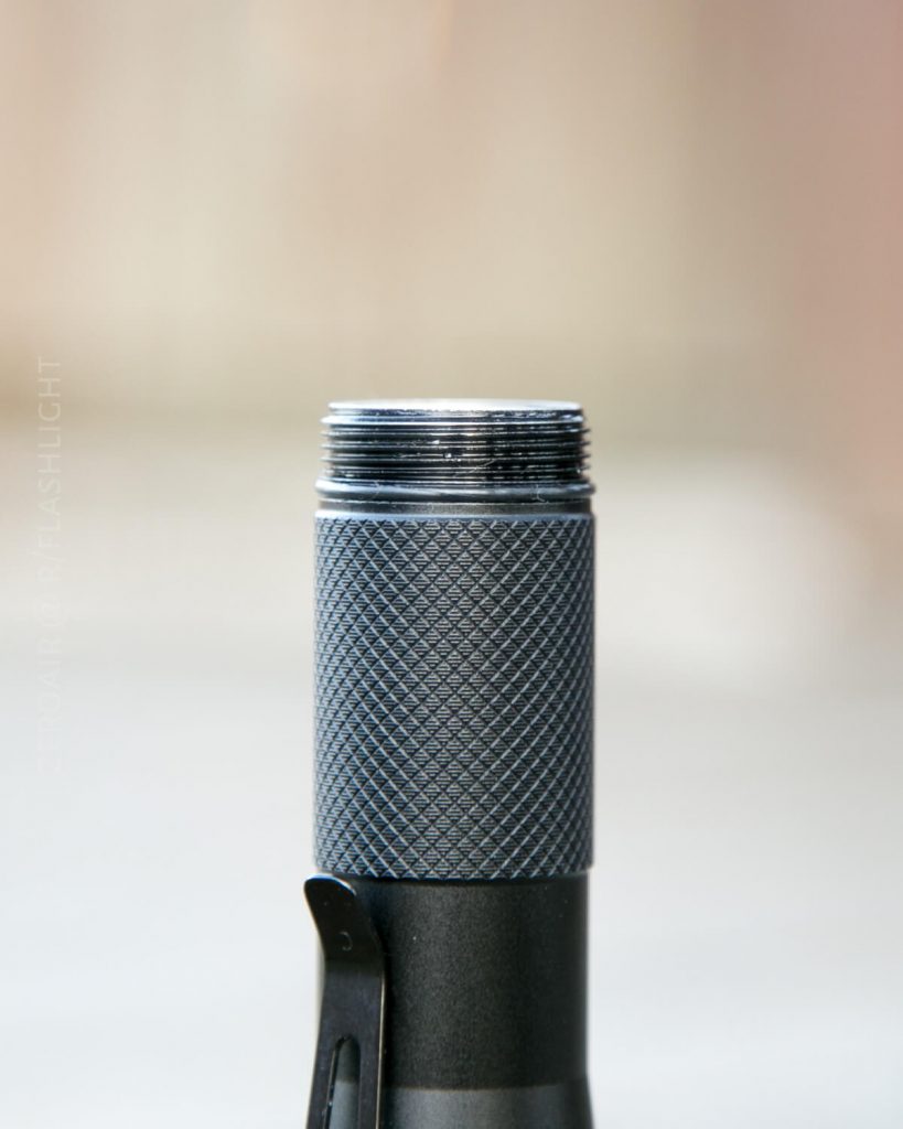 Close-up of the top part of a black metal flashlight with textured grip and visible screw threads, shown without its top or lens, against a blurred neutral background.
