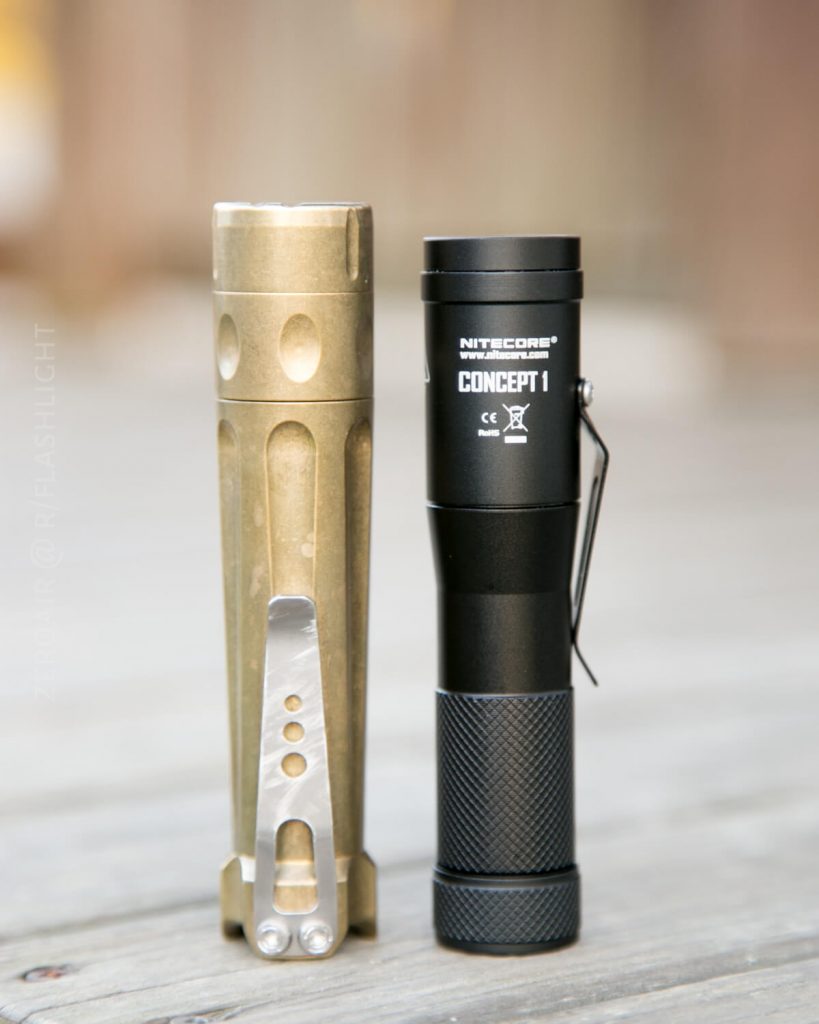 Two flashlights standing upright on a wooden surface. The left flashlight is metallic bronze with a grooved design and a silver pocket clip; the right is black with branding NITECORE CONCEPT 1 and a textured grip.