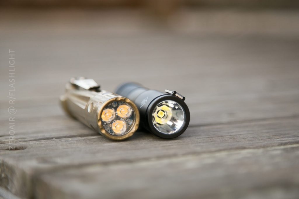 Two small flashlights are lying side by side on a wooden surface, one with three LED emitters and a brass finish, the other with a single LED emitter and a black finish.