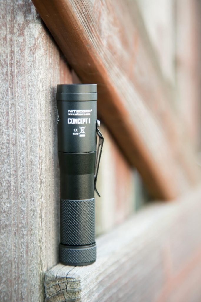 A black Nitecore Concept 1 flashlight stands upright against a wooden surface, with its side clip visible. The flashlight is positioned in natural daylight.