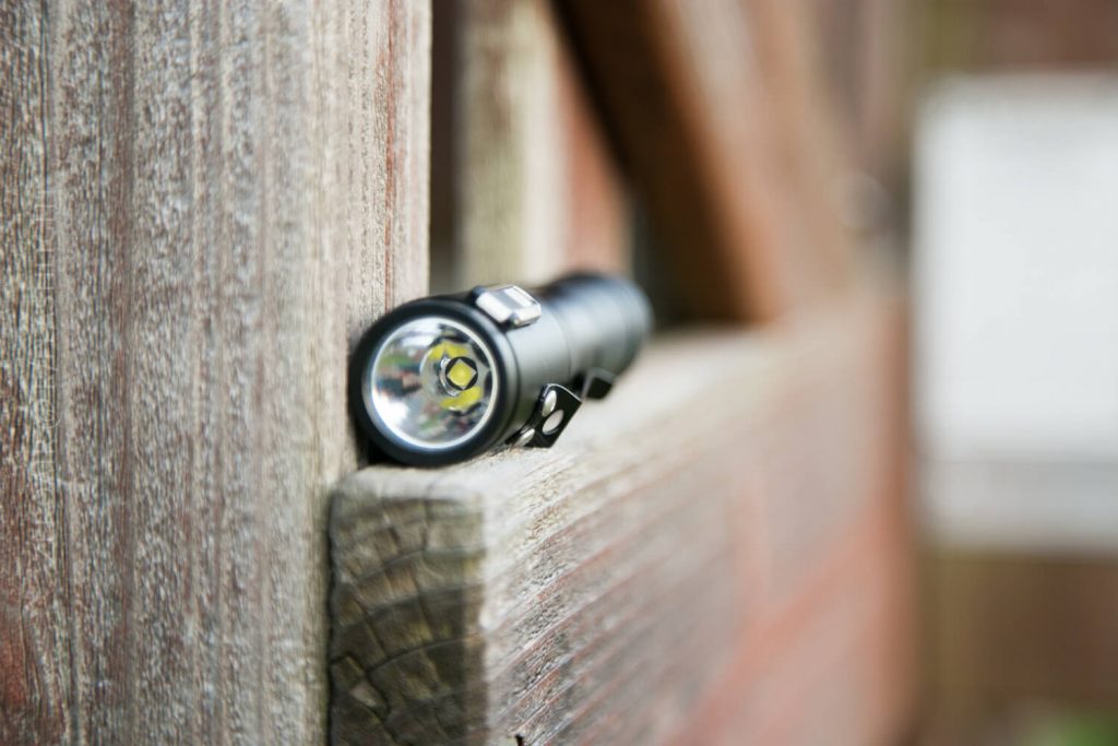 A black flashlight rests on a weathered wooden surface, with its lens facing the camera. The wood appears aged and has a rough texture. The background is out of focus.
