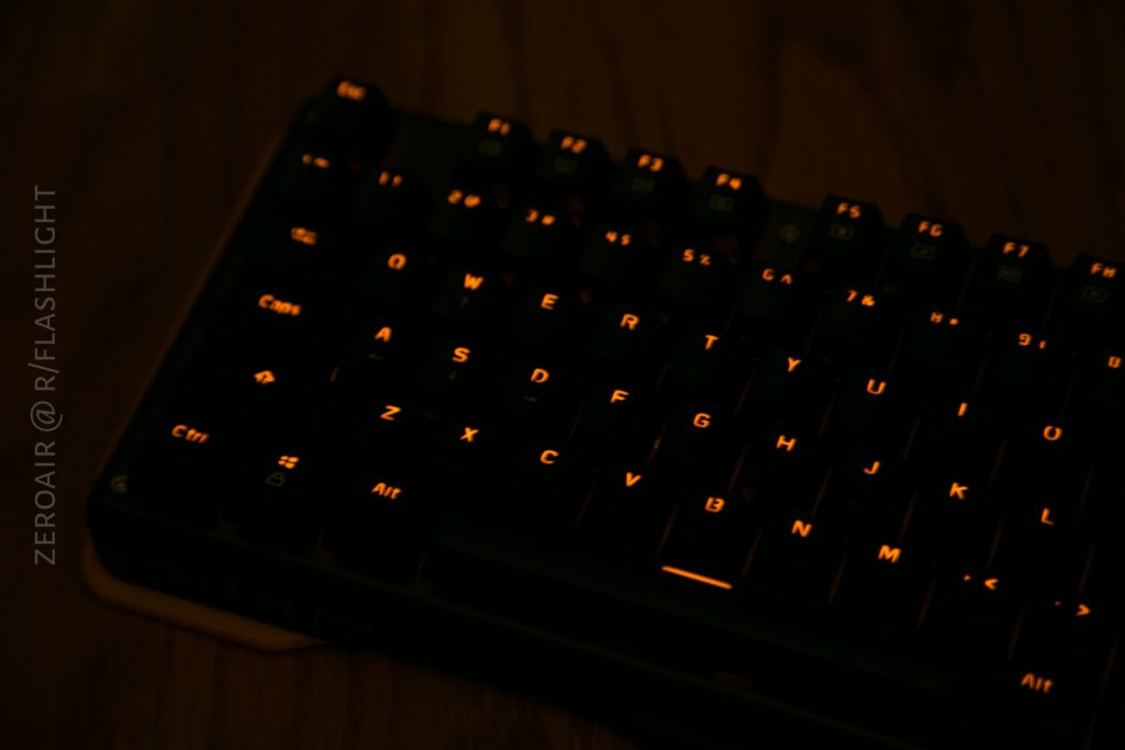A close-up of a black computer keyboard with orange backlit keys in a dark setting. The lighting highlights the letters and symbols on the keys. The words ZEROAIR @ R/FLASHLIGHT are visible on the left.