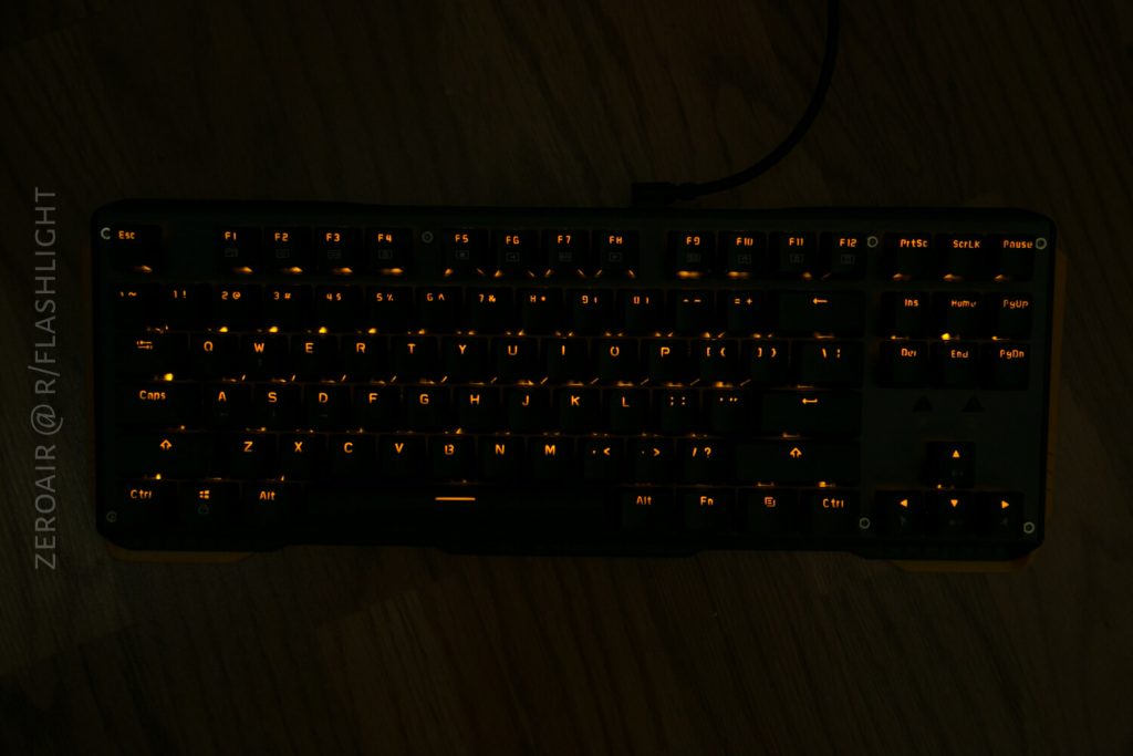 A black computer keyboard is shown with orange backlighting illuminating the keys. The background is a wooden surface. Text on the left side reads ZEROAIR @ R/FLASHLIGHT vertically.