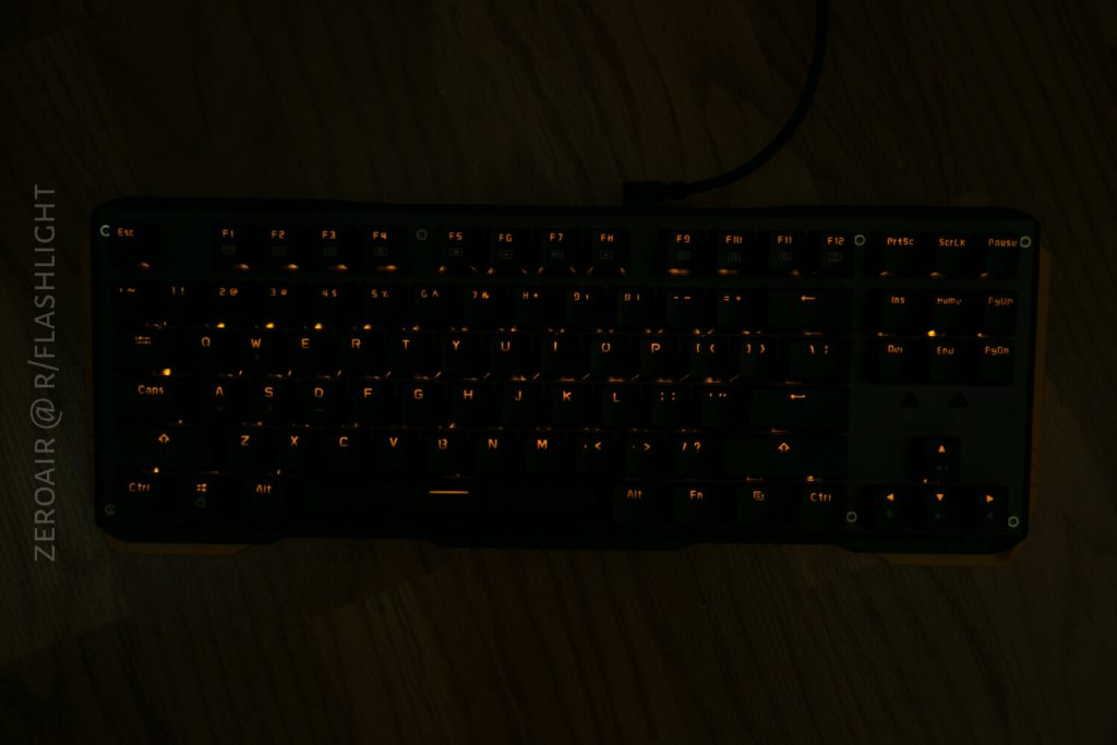 A black computer keyboard with orange backlit keys is shown on a wooden surface. The lighting is dim, making the key labels faintly visible. The text ZEROAIR @ R/FLASHLIGHT appears along the left side.