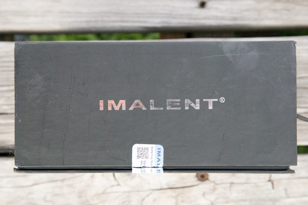 A black rectangular box with the brand name IMALENT printed in metallic letters on the lid, sitting on a wooden surface. A small sticker with QR codes and the word IMALENT is attached near the edge of the box.
