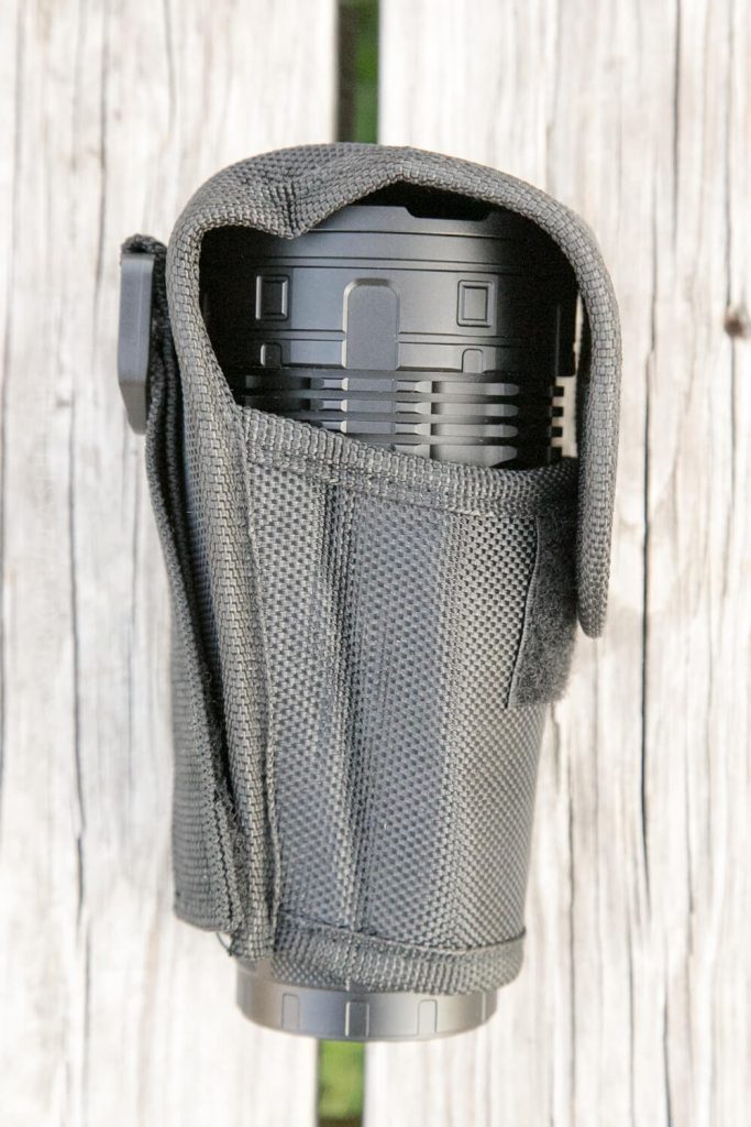 A black cylindrical object, possibly a flashlight or similar device, is partially enclosed in a textured black fabric case with a Velcro flap, placed on a weathered wooden surface.