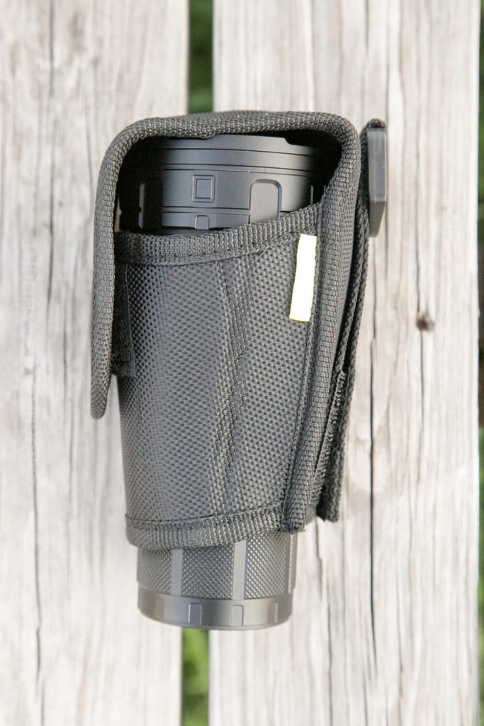 A black camera lens in a textured black lens pouch with a flap, resting vertically on a weathered wooden surface.