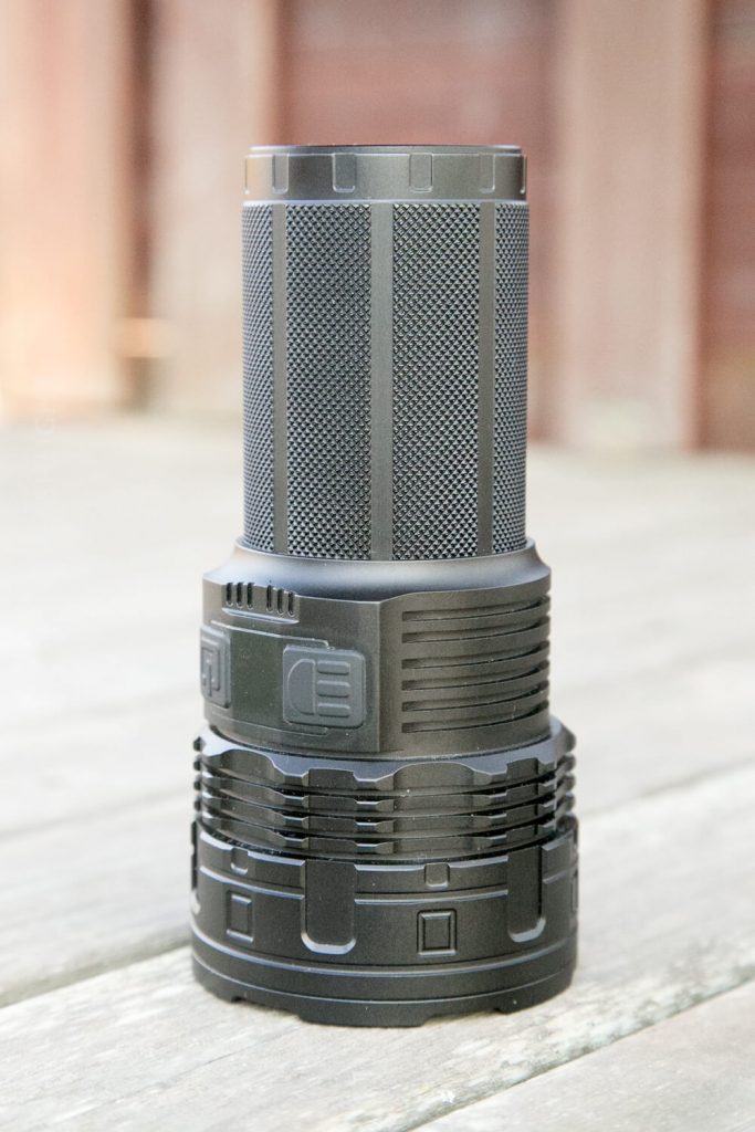 A large, black, cylindrical flashlight with textured grip and several buttons, standing upright on a wooden surface, with a blurred background.