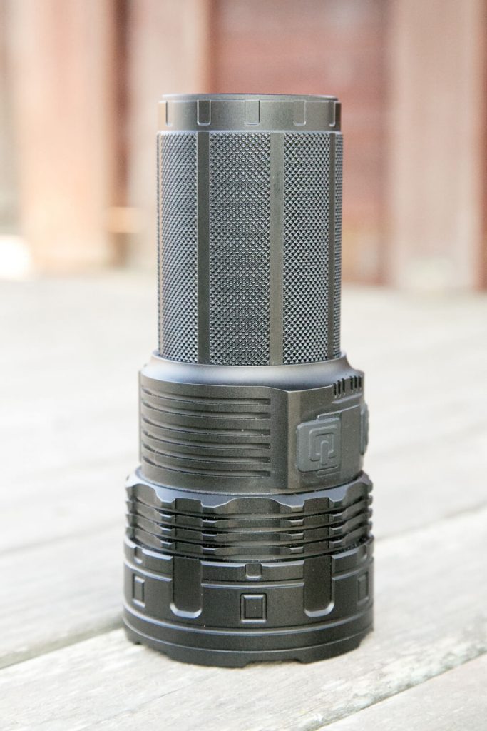 A black, cylindrical flashlight with a textured grip stands upright on a wooden surface. The background is blurred, showing hints of brown and beige tones.