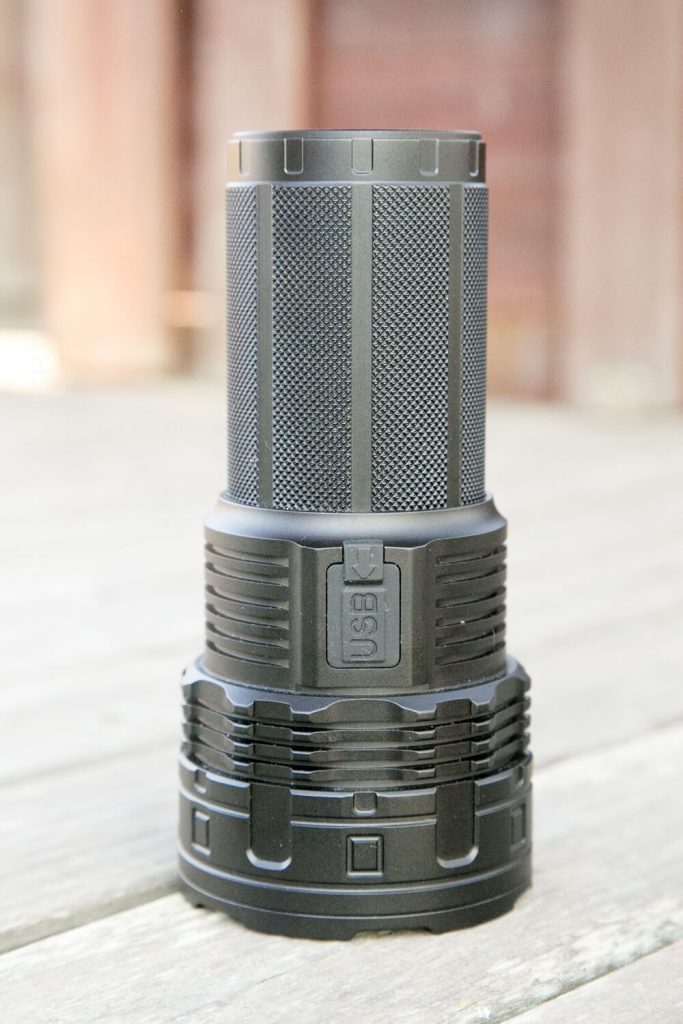 A black, cylindrical flashlight with a textured grip and a rectangular USB port cover sits upright on a wooden surface with a blurred background.