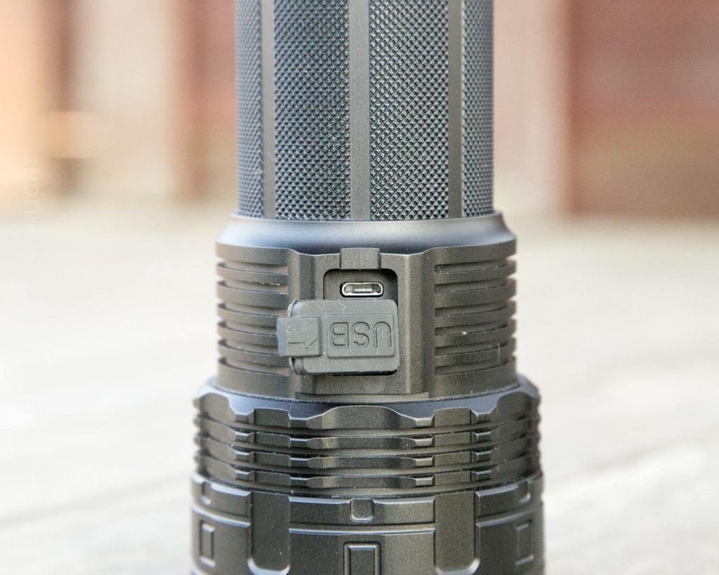 Close-up of a black flashlight showing a USB charging port covered by a rubber flap labeled USB. The background is blurred, and the focus is on the charging port area.