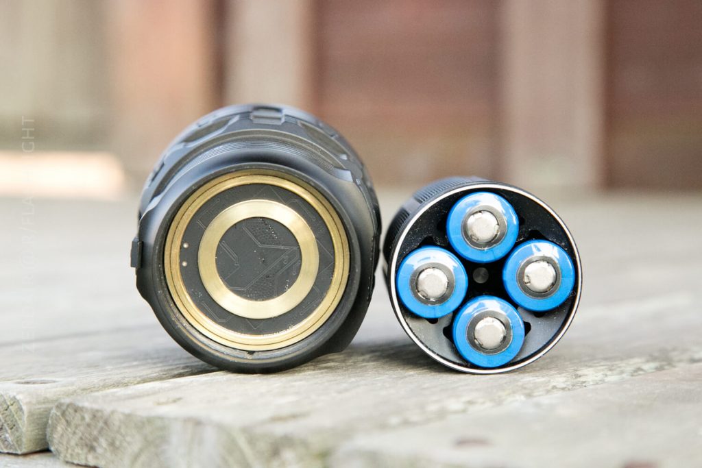A close-up of a flashlight with the tail cap removed, showing five blue cylindrical batteries installed in a circular configuration on a wooden surface.