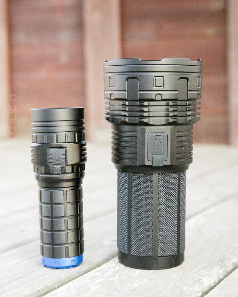 Two black flashlights, one smaller and one larger, stand upright on a wooden surface with a blurred wooden background. Both have cylindrical bodies and visible USB ports.