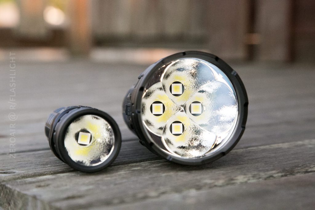 Two black flashlights are placed on a wooden surface. The larger flashlight has four LED bulbs visible inside its reflector, while the smaller flashlight has a single LED bulb. Both are facing forward.