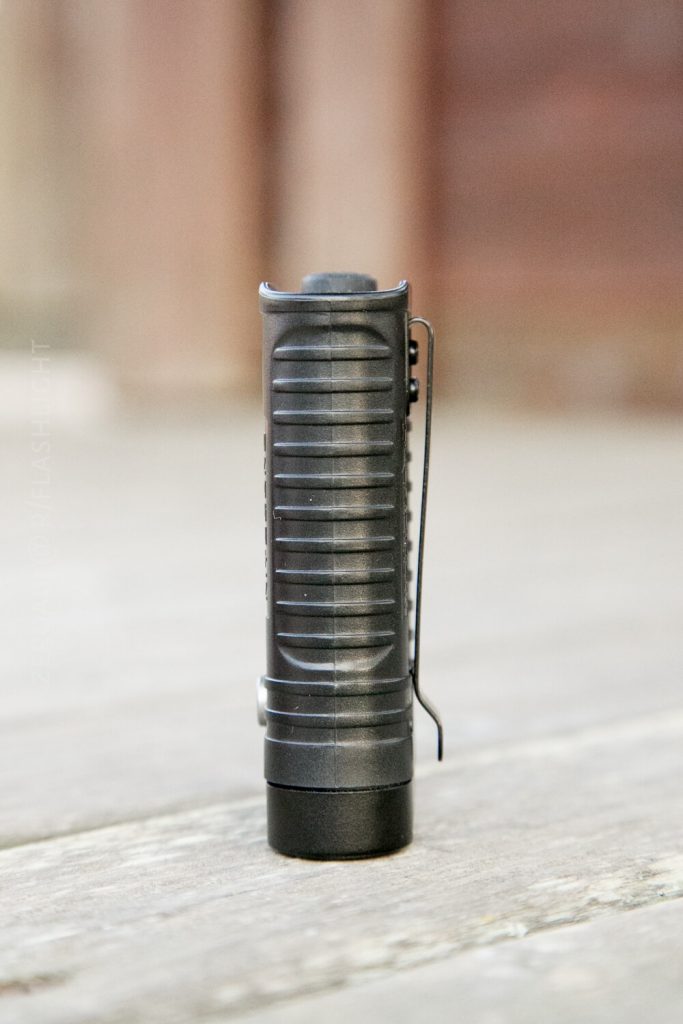 A small black flashlight with a ridged body and a pocket clip is standing upright on a wooden surface, with a blurred background.