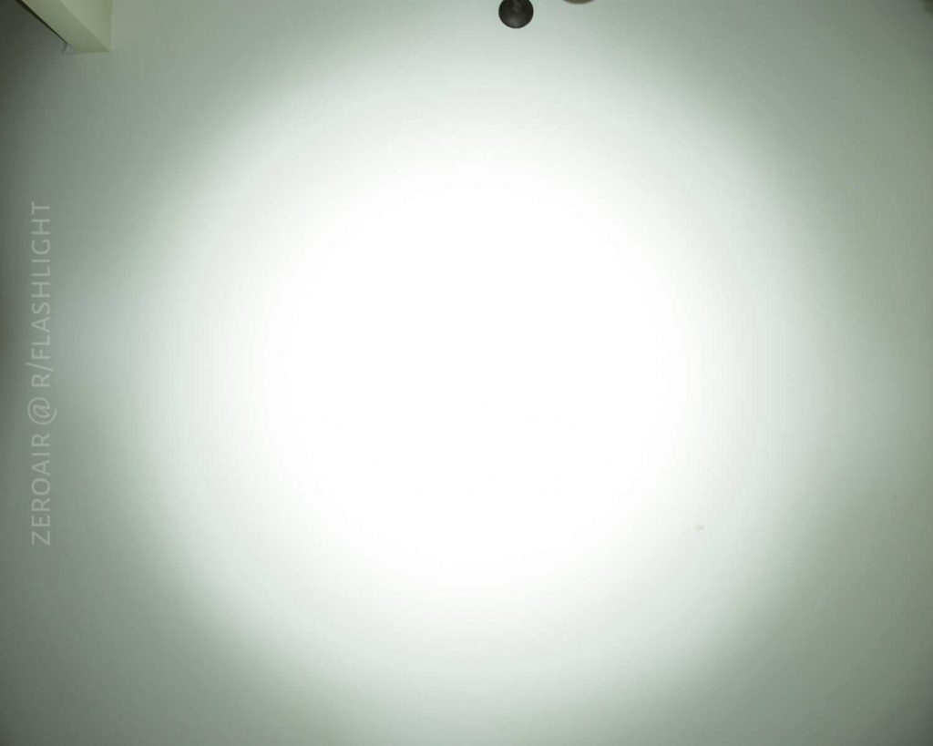A bright, circular beam of light is projected onto a plain white wall, with the text ZEROAIR @ R/FLASHLIGHT faintly visible on the left side. Two small dark circles appear near the top edge.
