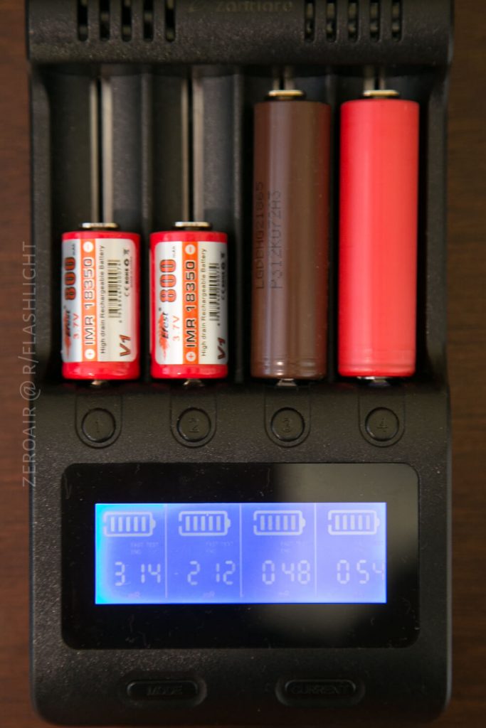 A battery charger holds four cylindrical batteries of different sizes and colors. An LCD screen below shows charging levels: 3.14, 2.12, 0.48, and 0.54 for each corresponding battery slot.