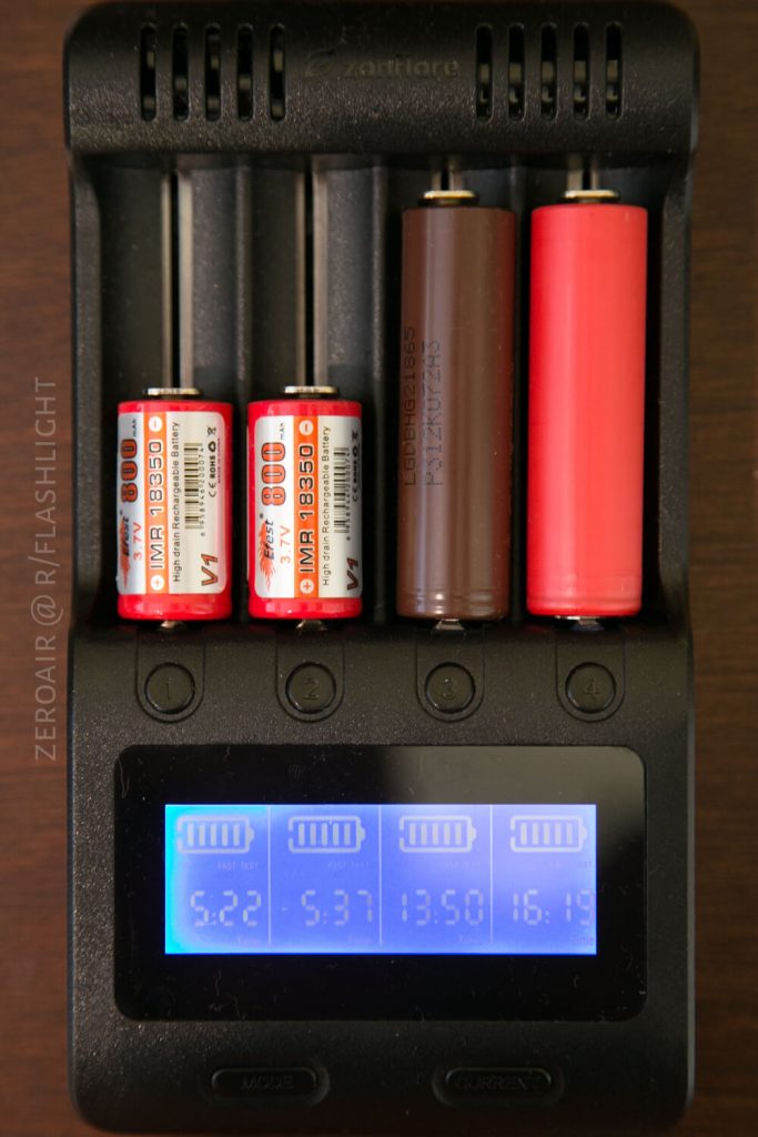 A battery charger holds four cylindrical batteries of different sizes. The digital display at the bottom shows four separate charge levels and corresponding voltages for each battery slot.