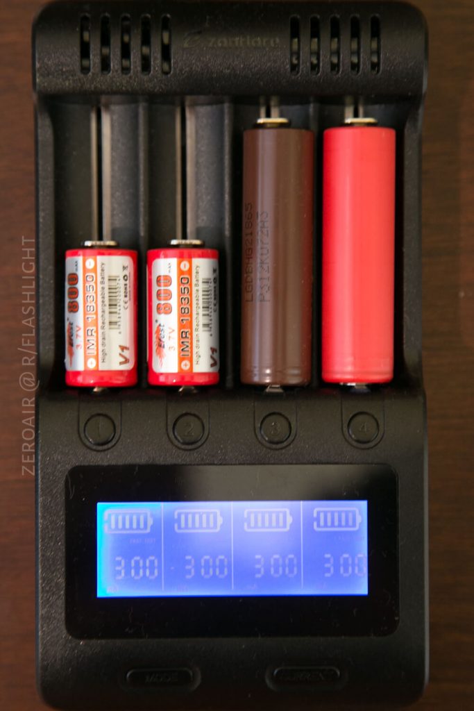 A battery charger holds four rechargeable batteries of different sizes and colors. The display screen shows the number 300 for each battery slot, indicating charging status. The charger rests on a wooden surface.
