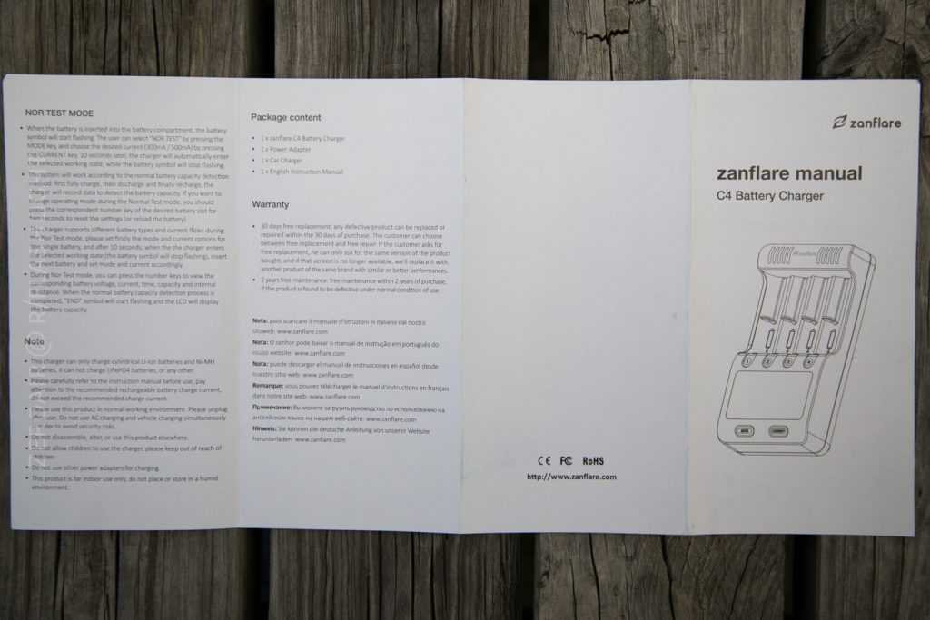 A Zanflare C4 Battery Charger manual is opened on a wooden surface, displaying instructions, package contents, warranty information, and a line drawing of the charger with batteries inserted.