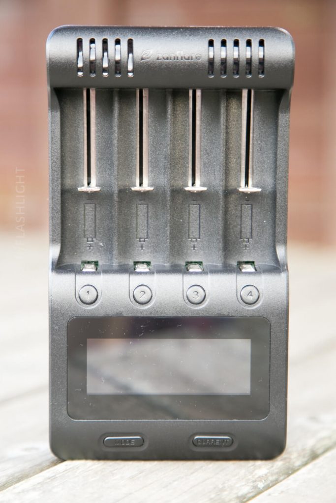 A black, four-slot battery charger with a digital display and numbered slot indicators, standing upright on a wooden surface. The brand name is visible at the top. The slots are currently empty.