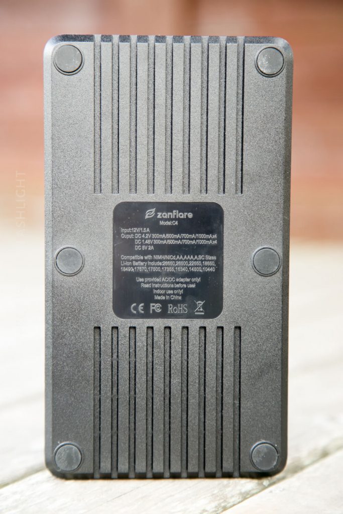A rectangular black electronic device, shown from the back, features vertical grooves and a central label with technical specifications and safety certifications. Four screws are visible at the corners.