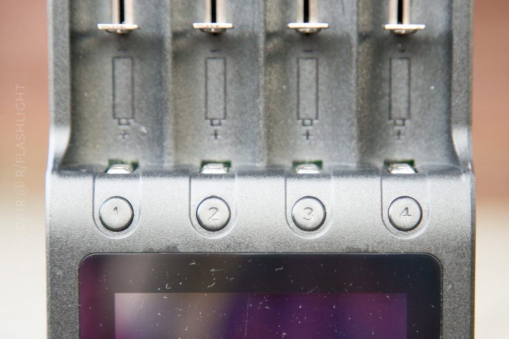 Close-up of a battery charger with four numbered charging slots, labeled 1 to 4. The slots display positive and negative symbols, and a portion of a digital display is visible at the bottom.