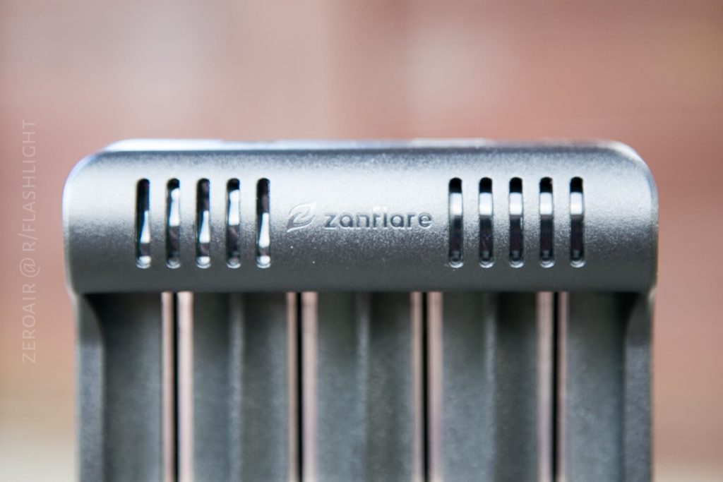 Close-up of a metallic object with vertical slots and the word zanflare engraved on it. The background is out of focus, and text ZEROAIR @ R/FLASHLIGHT appears along the left edge.