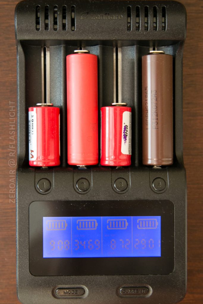 A battery charger holds four cylindrical batteries of different sizes and colors. The screen below displays individual voltage readings for each battery. The charger is placed on a dark brown surface.