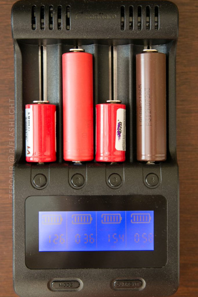 A black battery charger holds four cylindrical rechargeable batteries of different sizes and colors. A digital display at the bottom shows charge levels and numerical values for each battery slot.