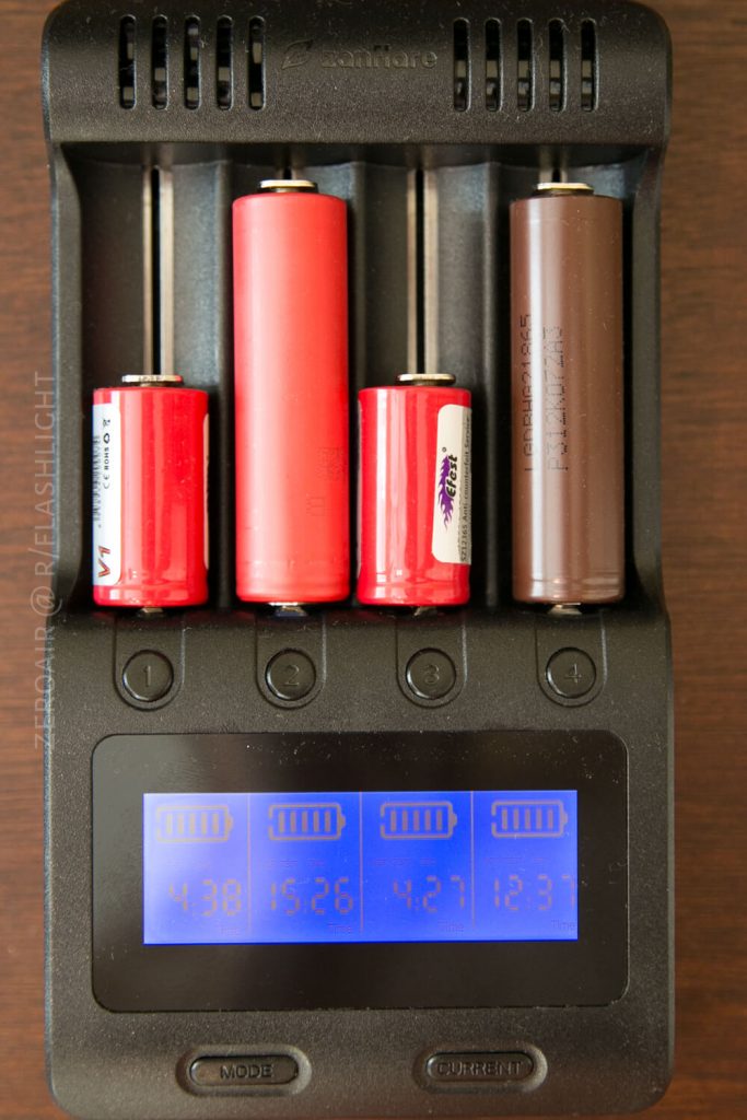 A black battery charger holds four cylindrical batteries of varying sizes and colors. The display shows voltage readings: 4.38, 15.26, 4.27, and 12.37. The charger rests on a wooden surface.