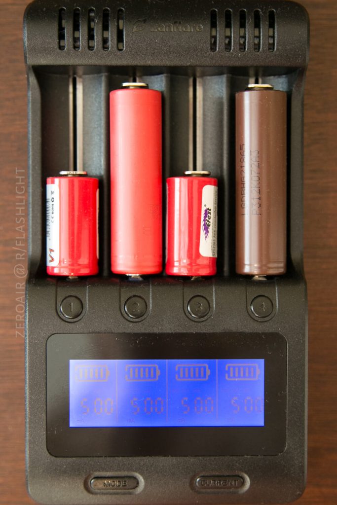 A battery charger holds four cylindrical batteries of varying sizes and colors, each slot showing a display reading 5.00 below the batteries on the digital screen. The background is a wooden surface.