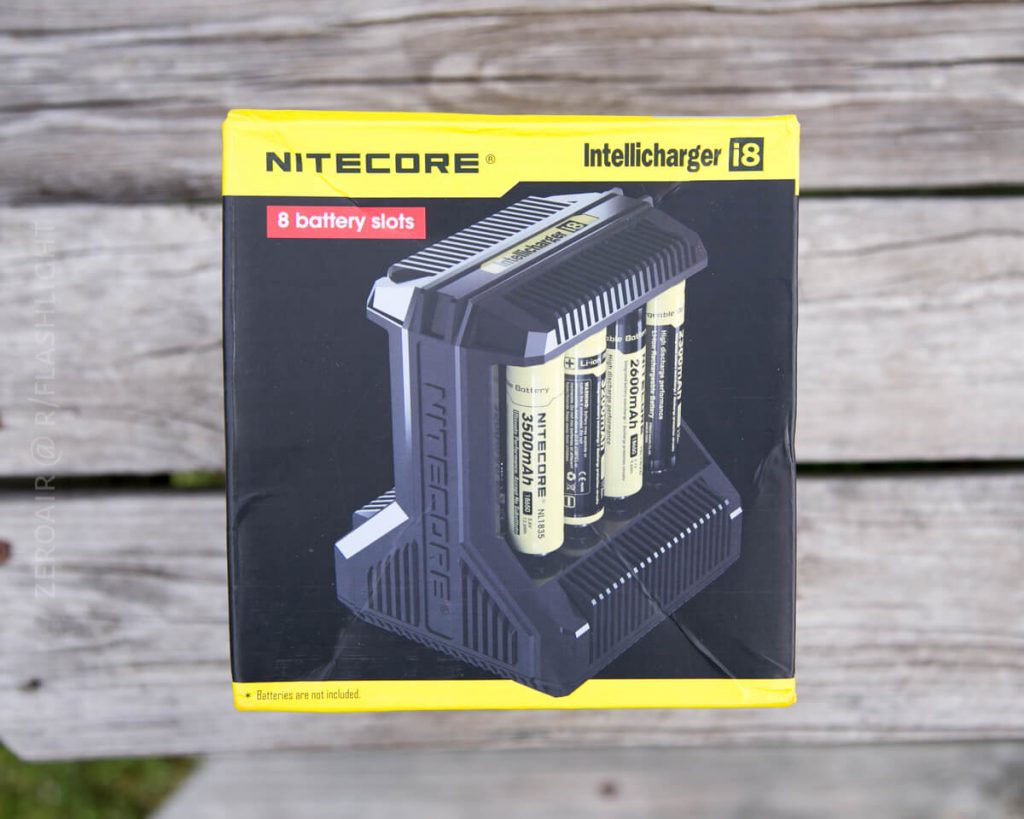 A box of a Nitecore Intellicharger i8 battery charger is shown on a wooden surface. The package highlights 8 battery slots and displays an image of the charger with batteries inserted.