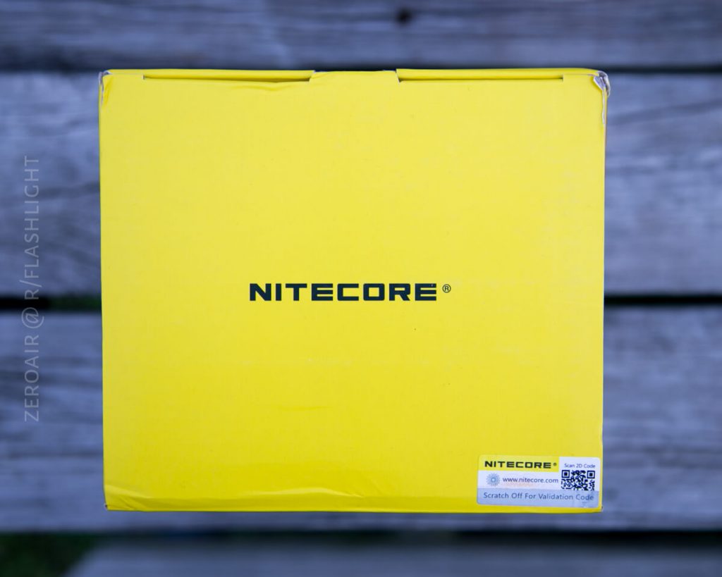A yellow Nitecore box with the brand name in black text is placed on a wooden surface. A white sticker with a QR code and validation code label is visible in the bottom right corner of the box.