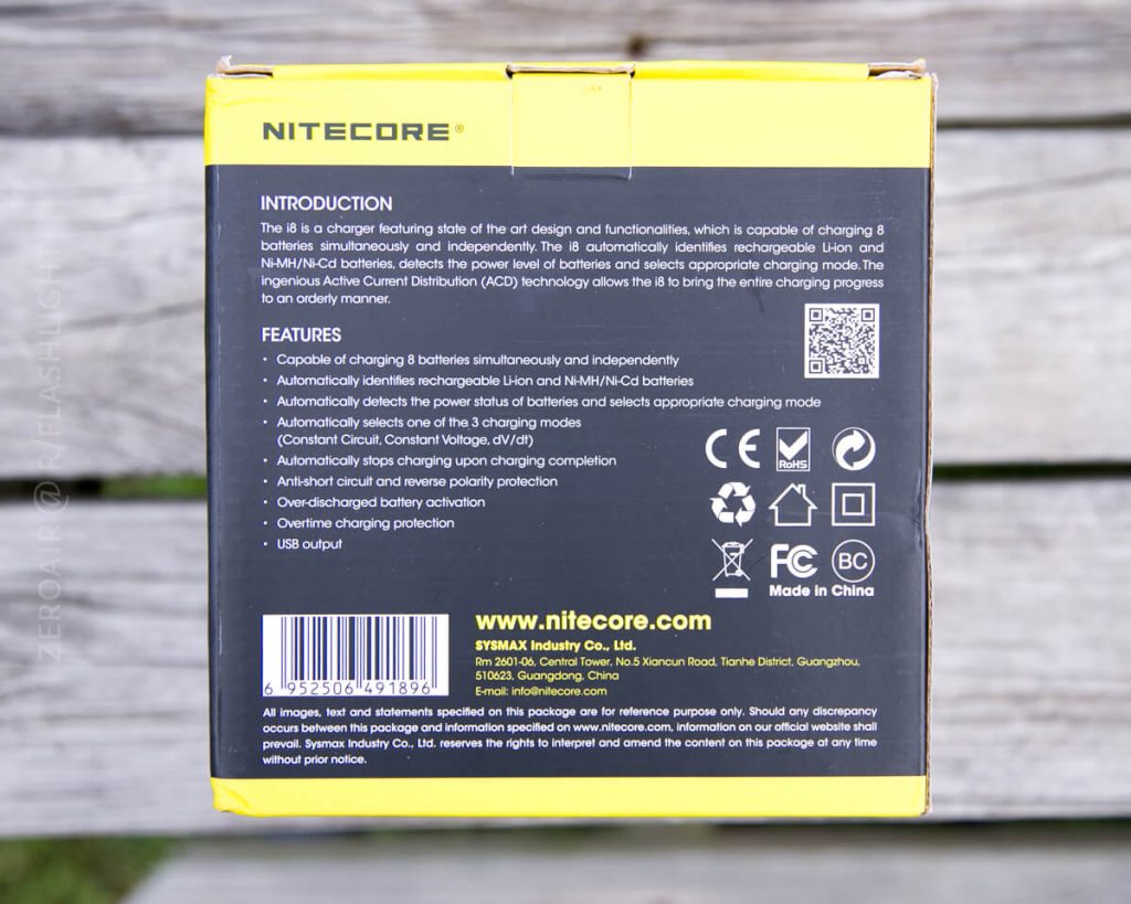 A yellow and black Nitecore battery charger box is shown. The back lists product features and specifications in white text, with icons for safety certifications and a barcode at the bottom. The box is placed on a wooden surface.