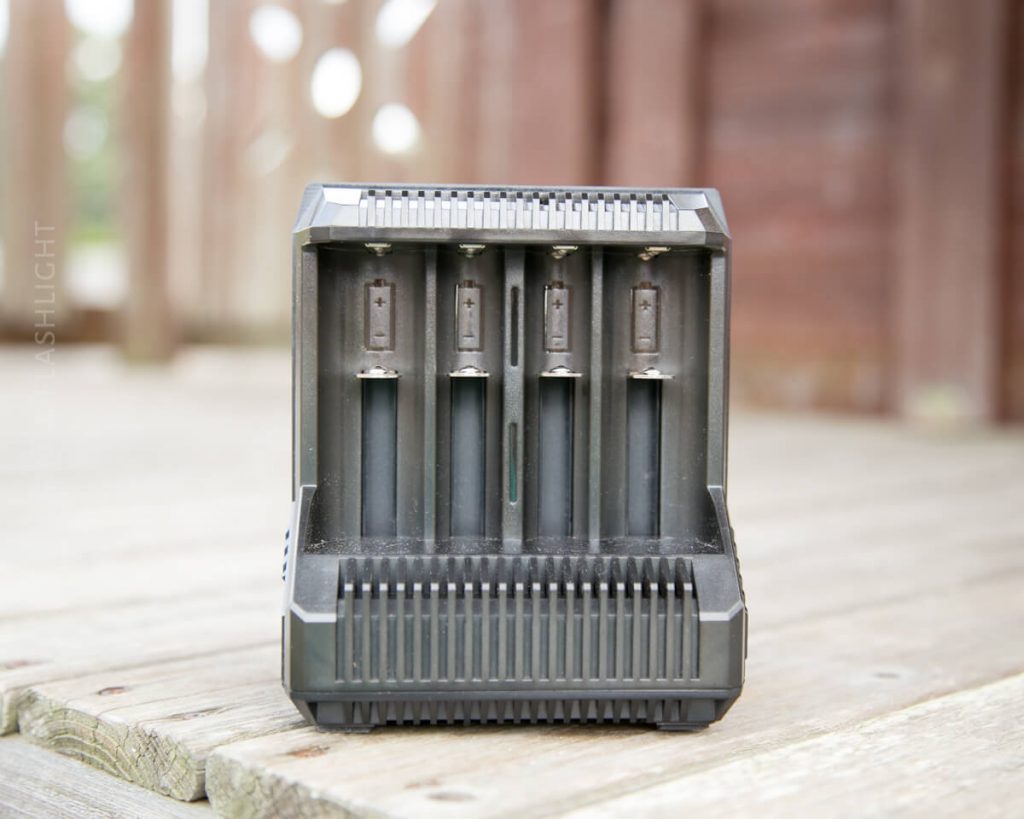 A black battery charger with four empty slots is placed on a wooden surface outdoors, with a blurred wooden structure in the background.