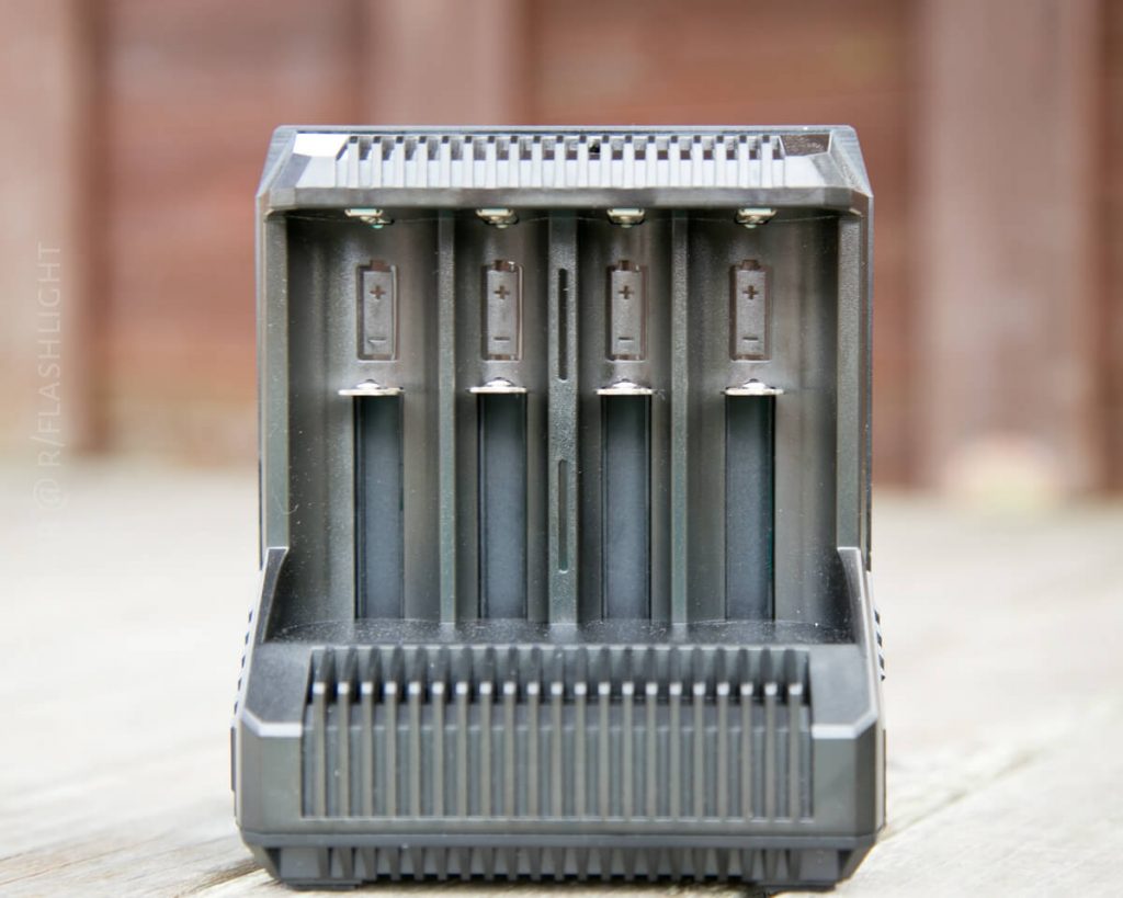 A close-up of a black battery charger with four vertical charging slots, each marked with plus and minus symbols. The charger is placed on a wooden surface with a blurred brown background.