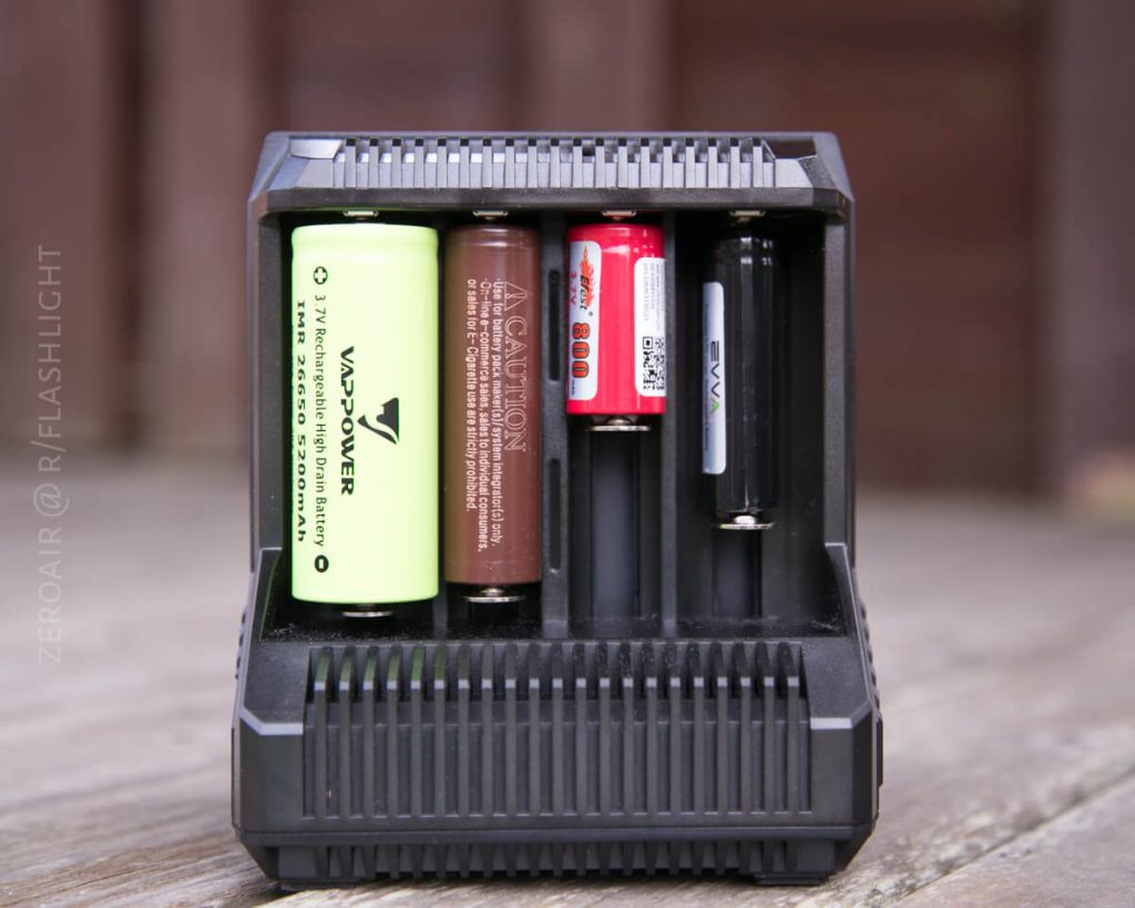 A black battery charger is placed on a wooden surface, holding four different types of batteries in separate charging slots. The background is blurred.