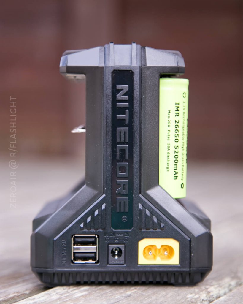 A Nitecore battery charger holding a green IMR 26500 5200mAh battery, showing USB, DC input, and output ports on a wooden surface. The Nitecore logo is visible on the front of the device.