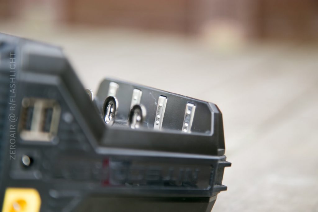 A close-up of a black battery charger with four charging slots, some containing metal contacts and one with an AA battery. The background is blurred.