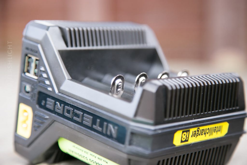 A close-up view of a Nitecore battery charger with an inserted green and yellow AA rechargeable battery labeled Intelligent 1.8V. The charger is black with visible slots and metal contacts.