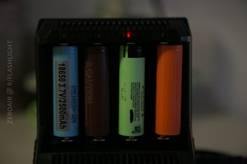 A close-up of a battery charger holding four cylindrical batteries of different colors and brands, with a small red indicator light visible at the top of the charger.