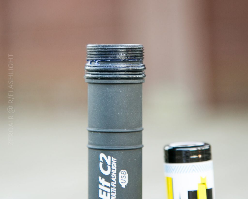 A close-up of the top of a black Elf C2 multi-flashlight with visible threading, next to a partially visible battery standing upright on a blurred background.
