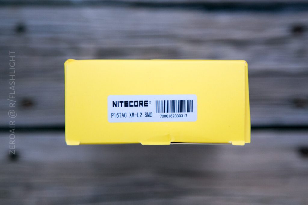 A close-up of the top of a yellow box labeled NITECORE with a barcode and the text P16TAC XM-L2 SMO 700617830317. The background is a wooden surface.
