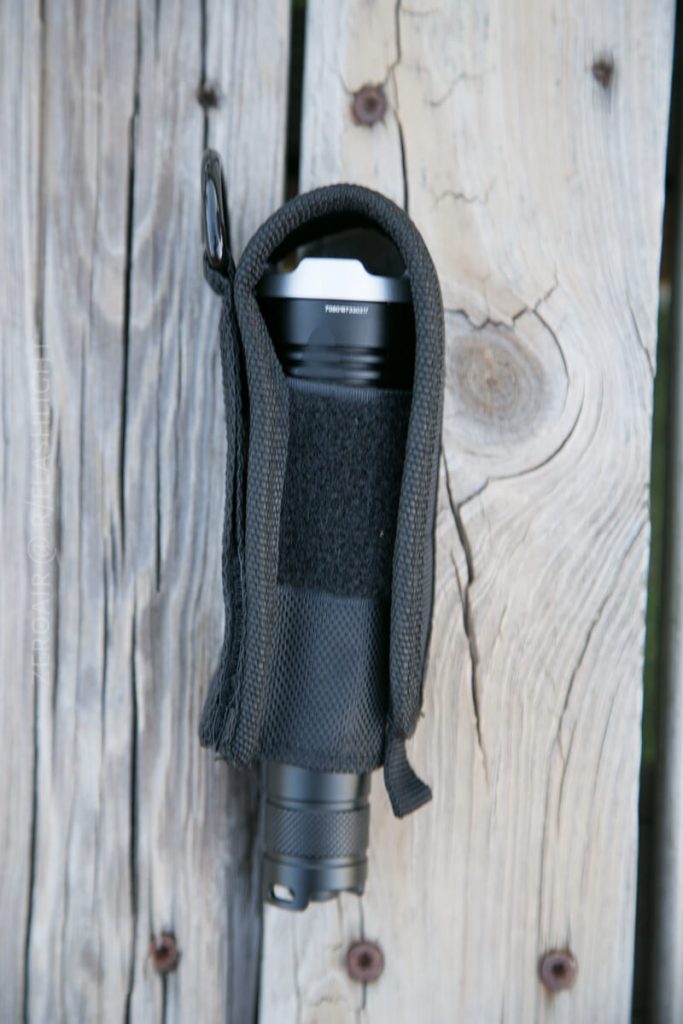 A black flashlight in a nylon carrying case is placed vertically on a weathered wooden surface with visible knots and grain patterns.