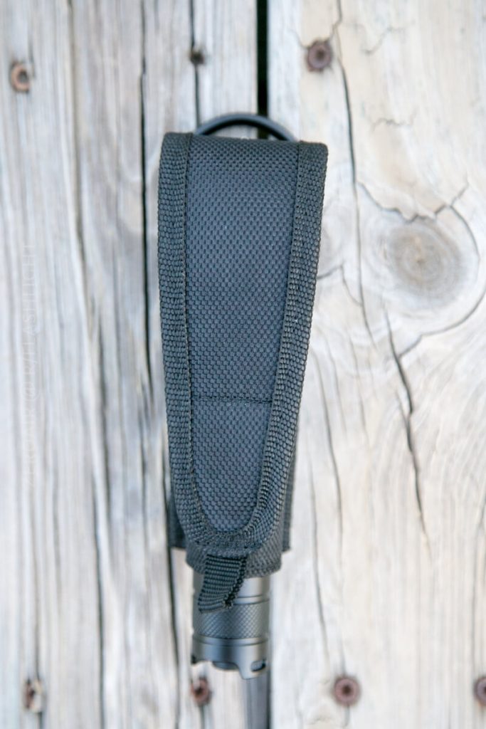 A black textured nylon flashlight holster with a flashlight partially visible at the bottom, resting vertically on a weathered wooden surface with visible knots and grain.