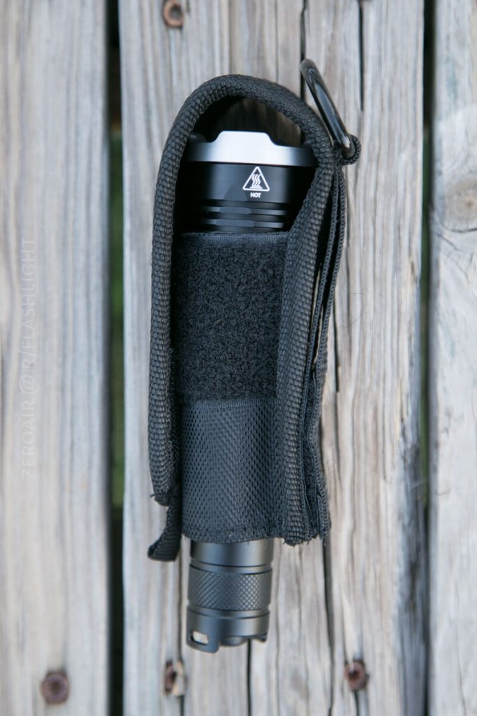 A black flashlight in a nylon holster is attached to a weathered wooden surface. The holster is partially open, showing the flashlight head and textured grip.