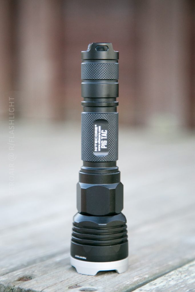 A black Nitecore P16 TAC tactical flashlight is standing upright on a wooden surface, with a blurred background. The flashlight features a textured grip and a silver bezel at the base.