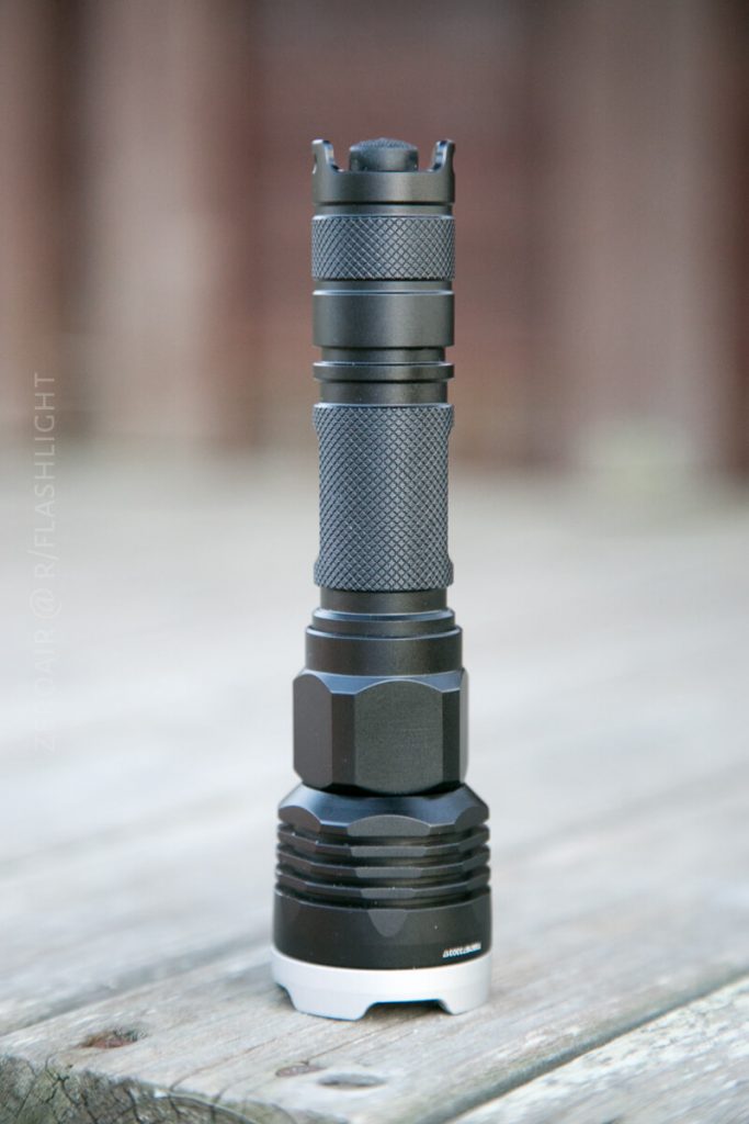 A black tactical flashlight stands upright on a wooden surface. The flashlight has a textured grip and a wide base, with a blurred brown background.