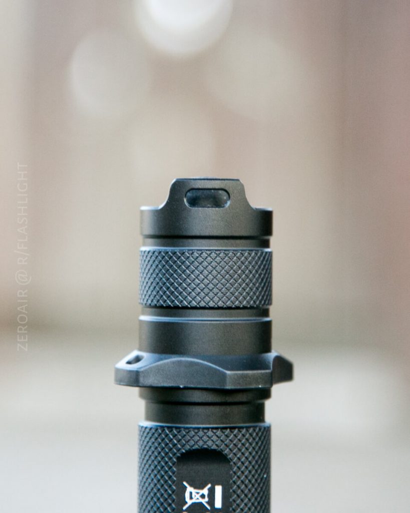 Close-up of the top section of a black metal flashlight with textured grip and a slot near the top, against a blurry neutral background. The words ZEROAIR @ R/FLASHLIGHT are faintly visible on the left side.